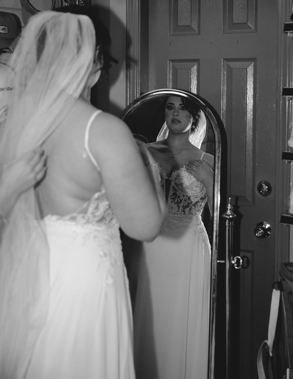 Black and white photo of bride in lace wedding dress viewing her reflection in a full-length mirror.