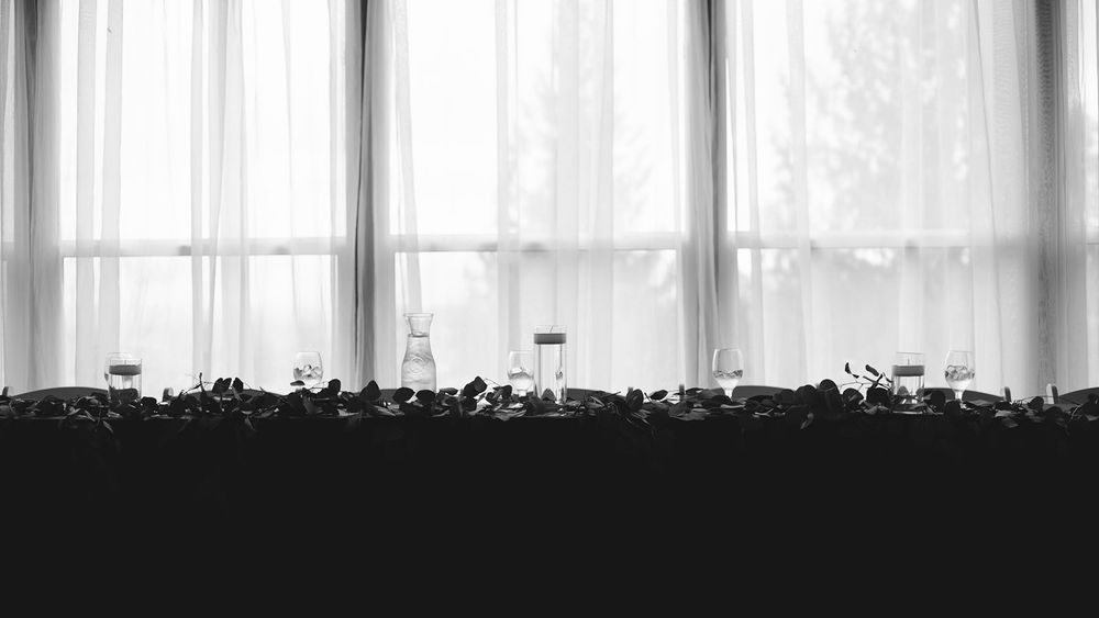 Black and white wide shot of wedding reception table setup against sheer curtained windows.