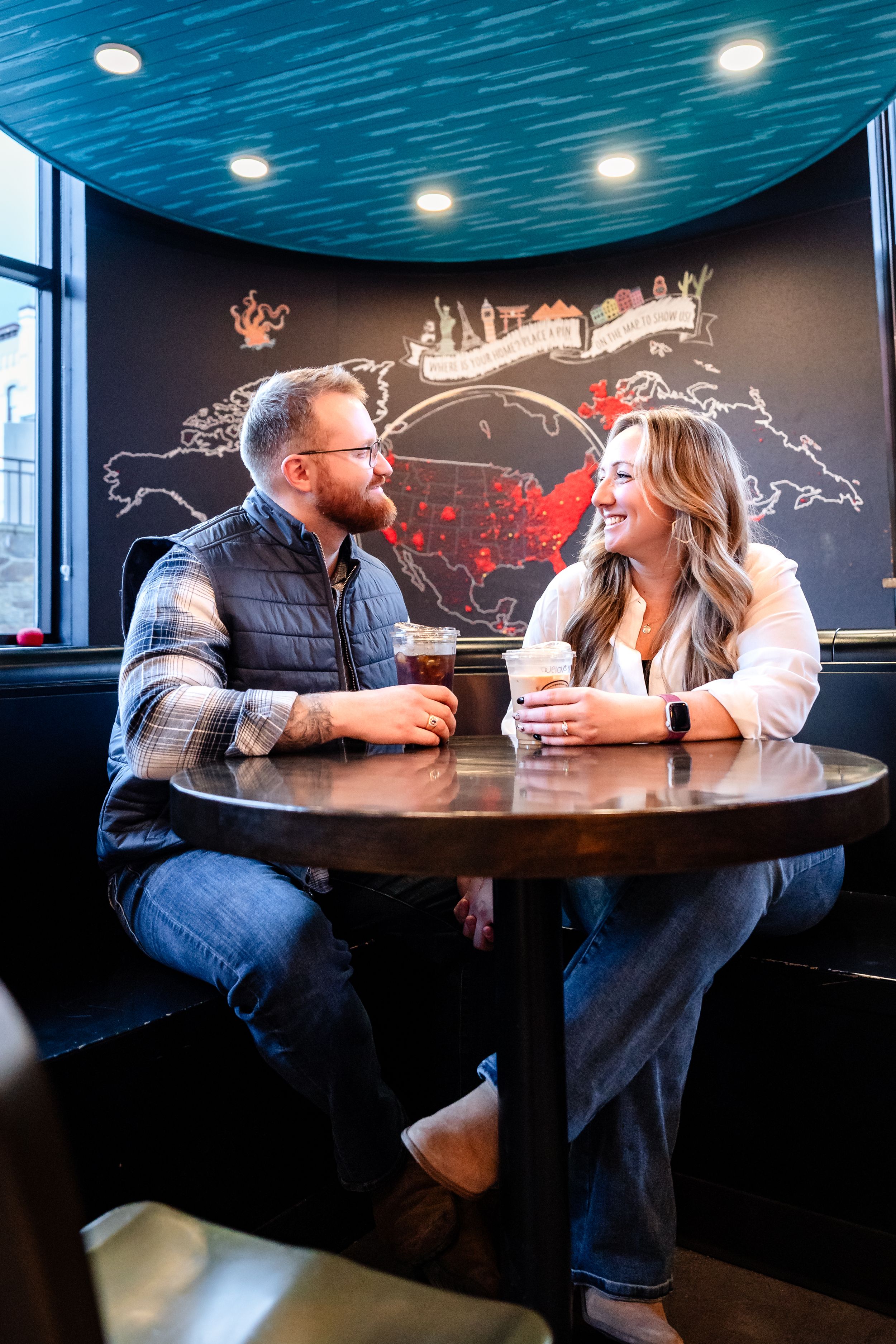 A couple enjoys drinks and conversation at a cozy restaurant booth with blue lighting and world map decor.