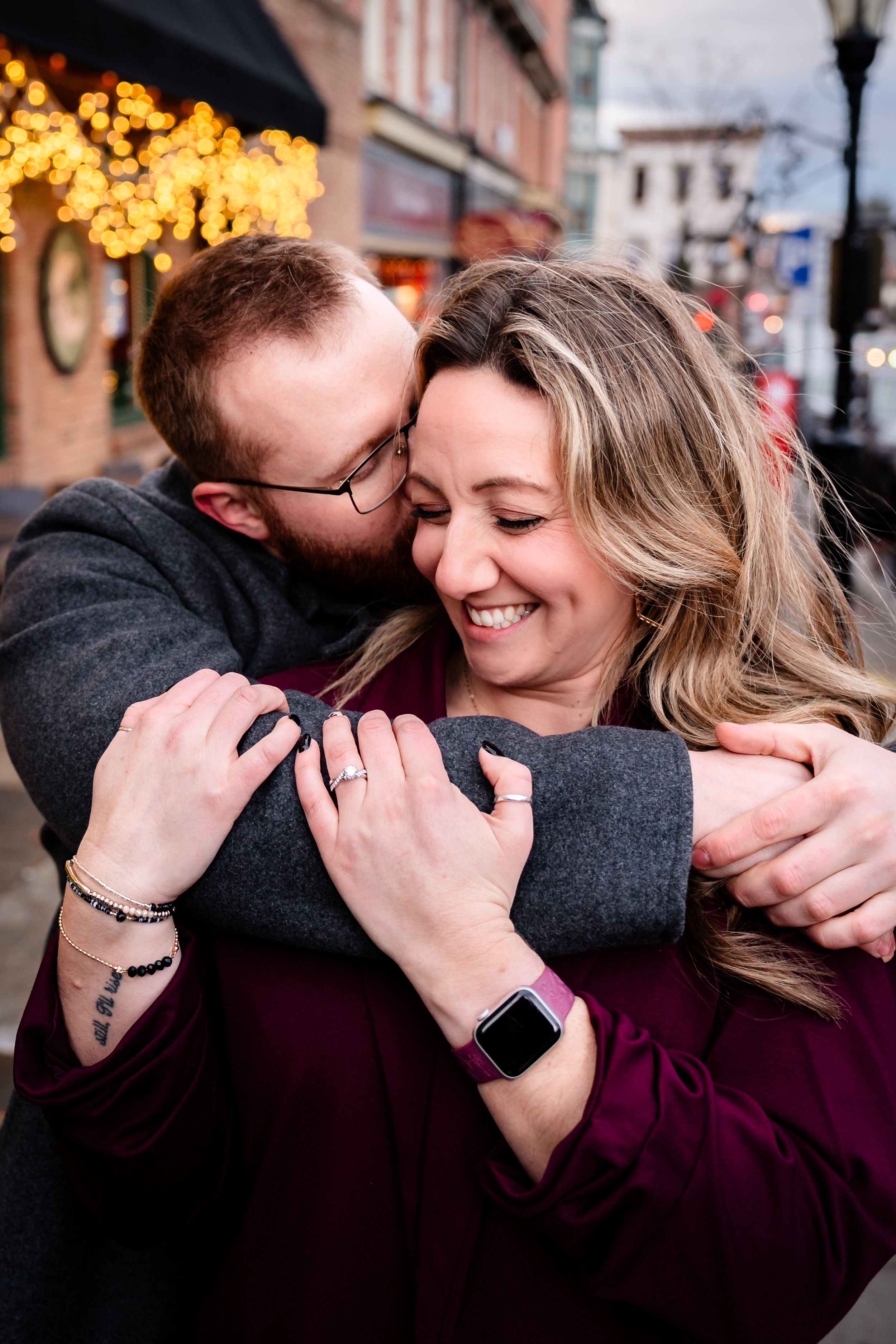 A couple embraces and laughs together against a backdrop of warm city lights at dusk.