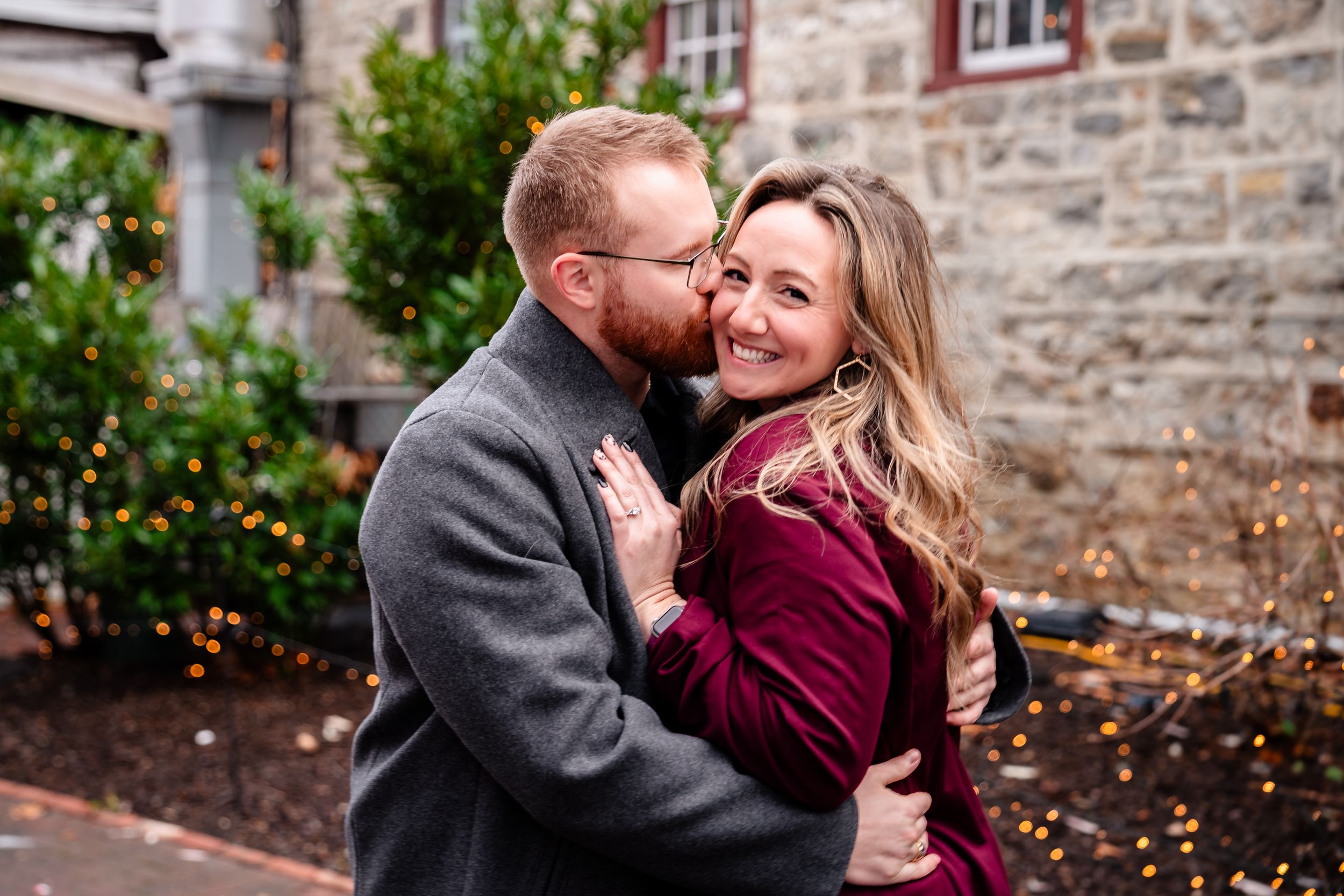 A romantic couple embraces outdoors against a stone wall on a fall day, with one wearing grey and the other in burgundy.