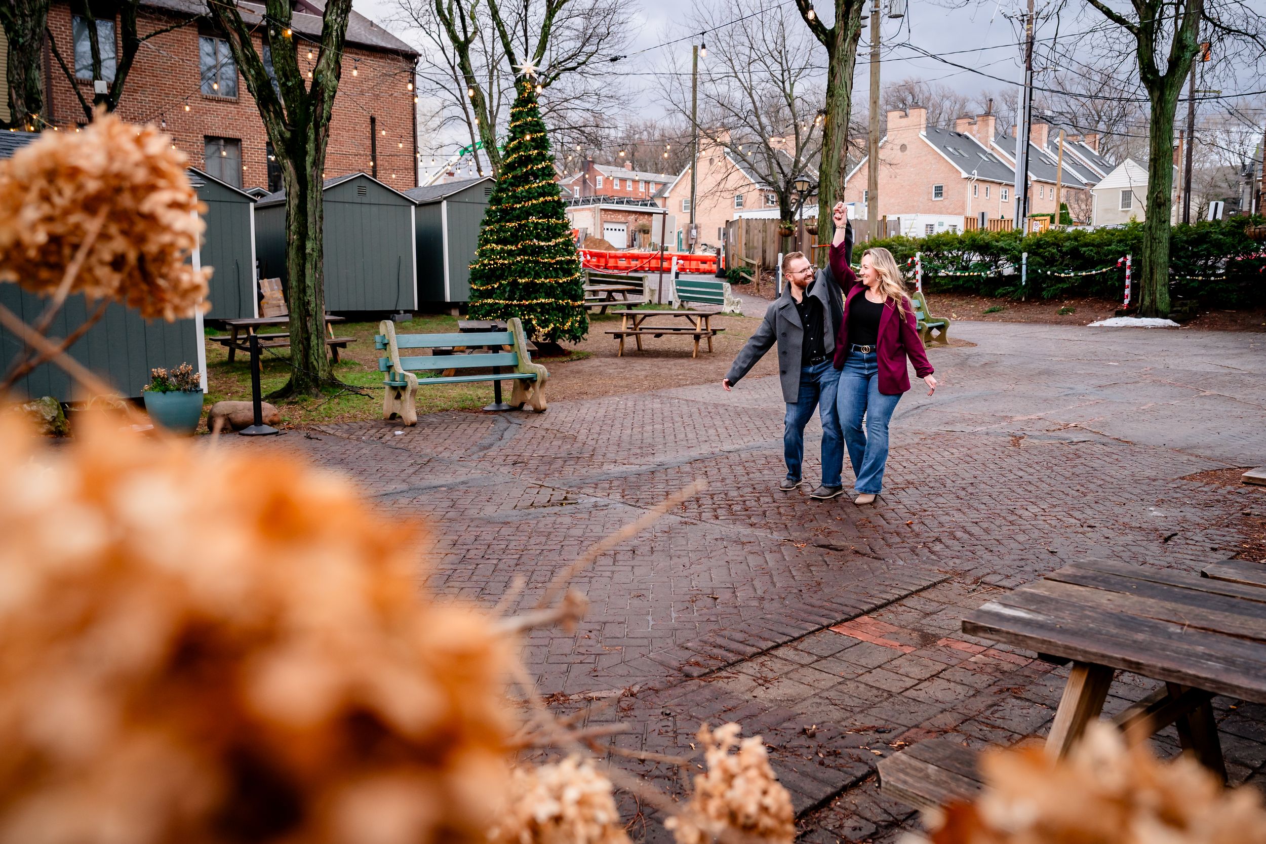 A couple walks hand in hand along a brick pathway in a quaint town setting during autumn.
