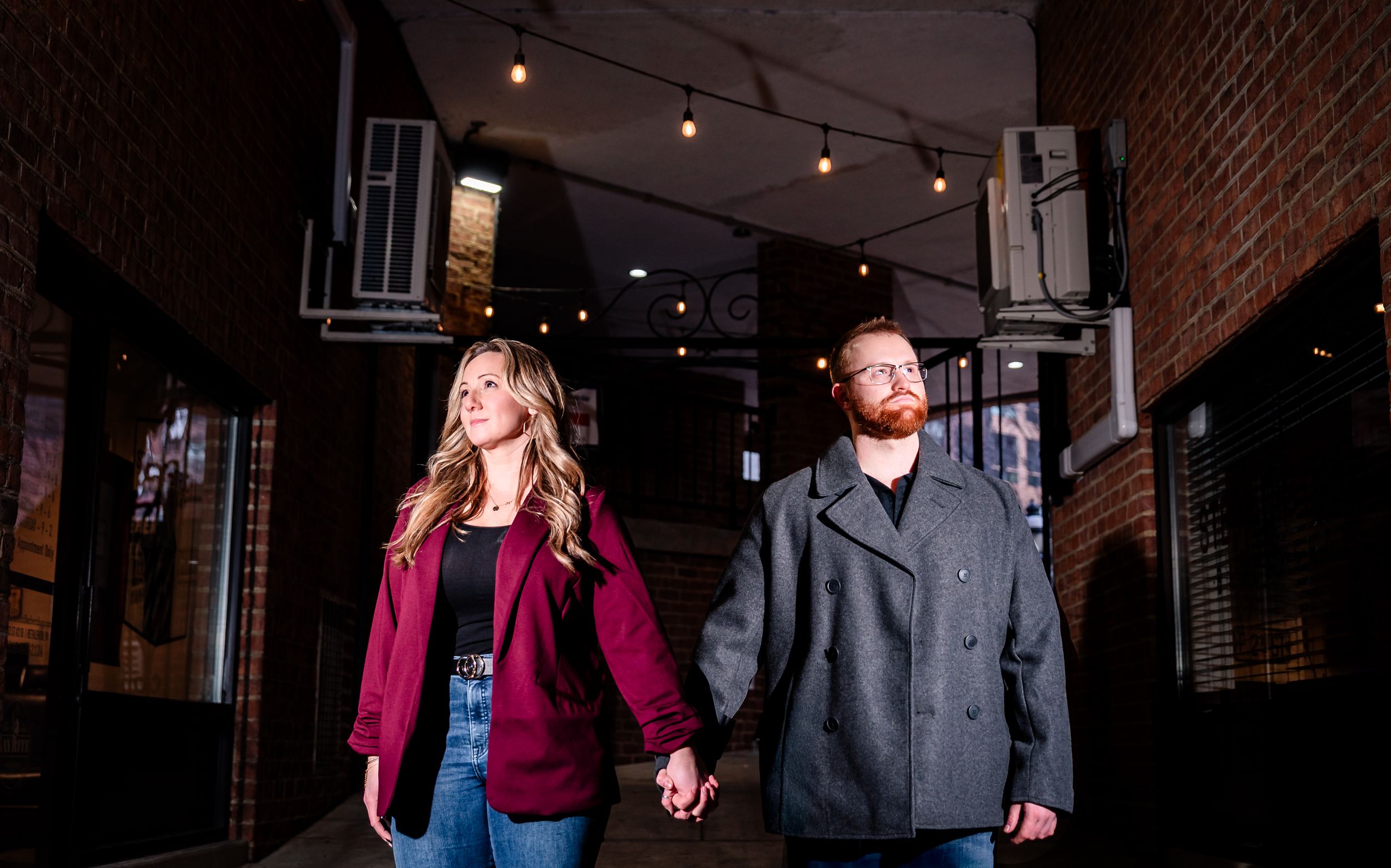 A couple stands close together under warm lighting against a rustic brick wall backdrop.