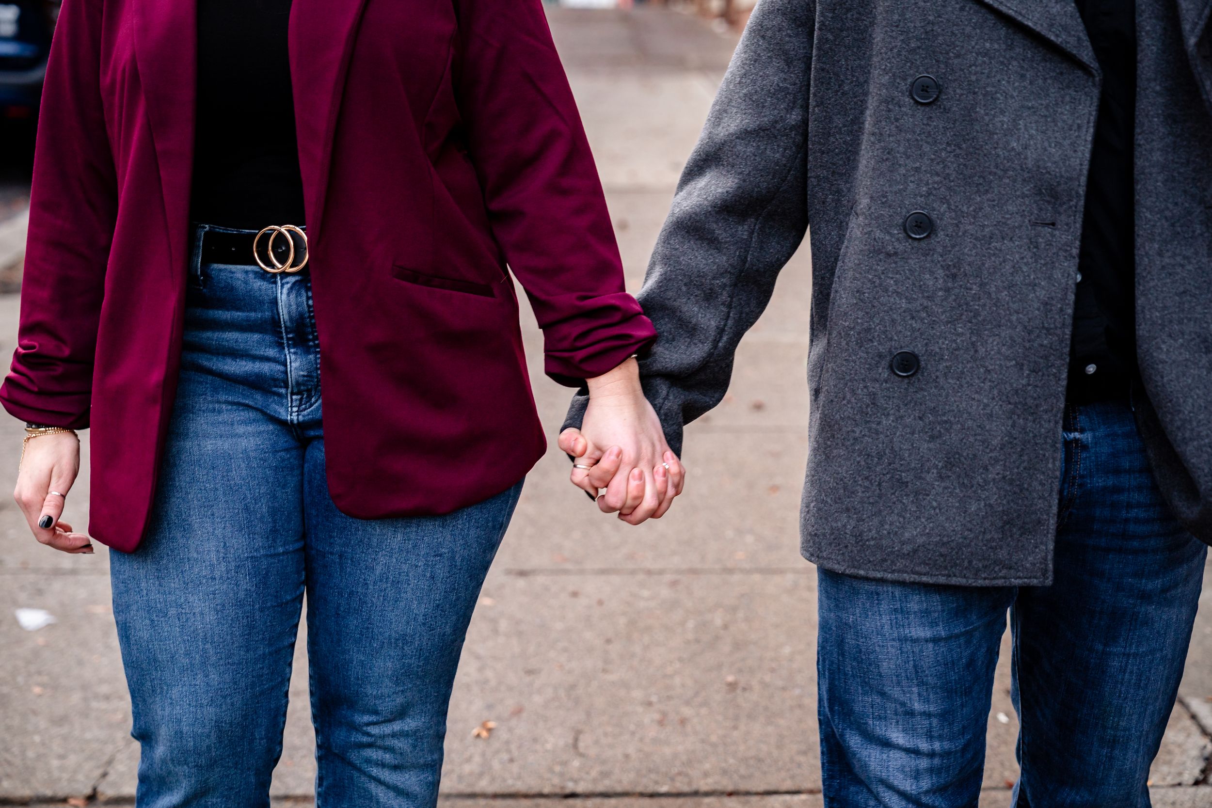 A couple in burgundy blazers and jeans hold hands while walking together on a street.