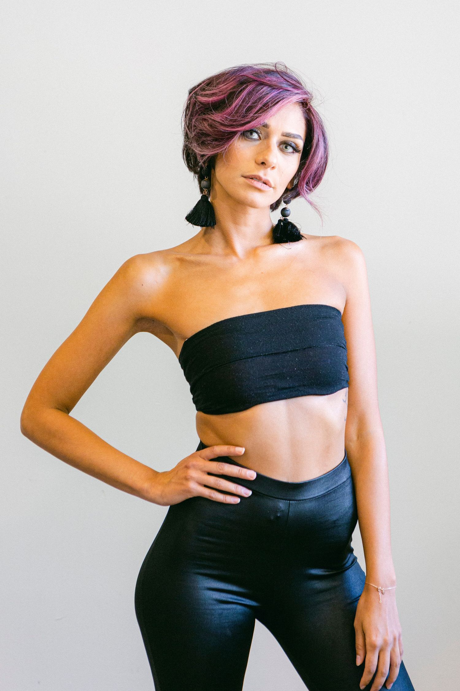 Athletic figure in black bandeau top and high-waisted pants poses with purple-tinted hair against light background.