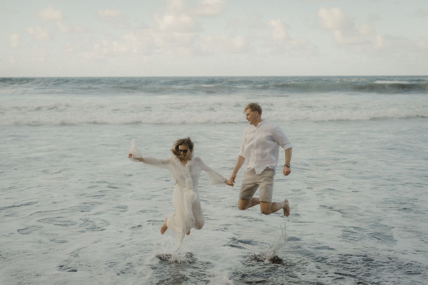 A couple in white outfits playfully dances and splashes in the ocean waves at sunset.