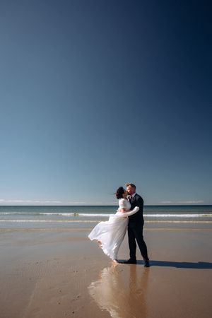 A couple in wedding attire shares a romantic moment on a serene beach with blue skies and calm waters.