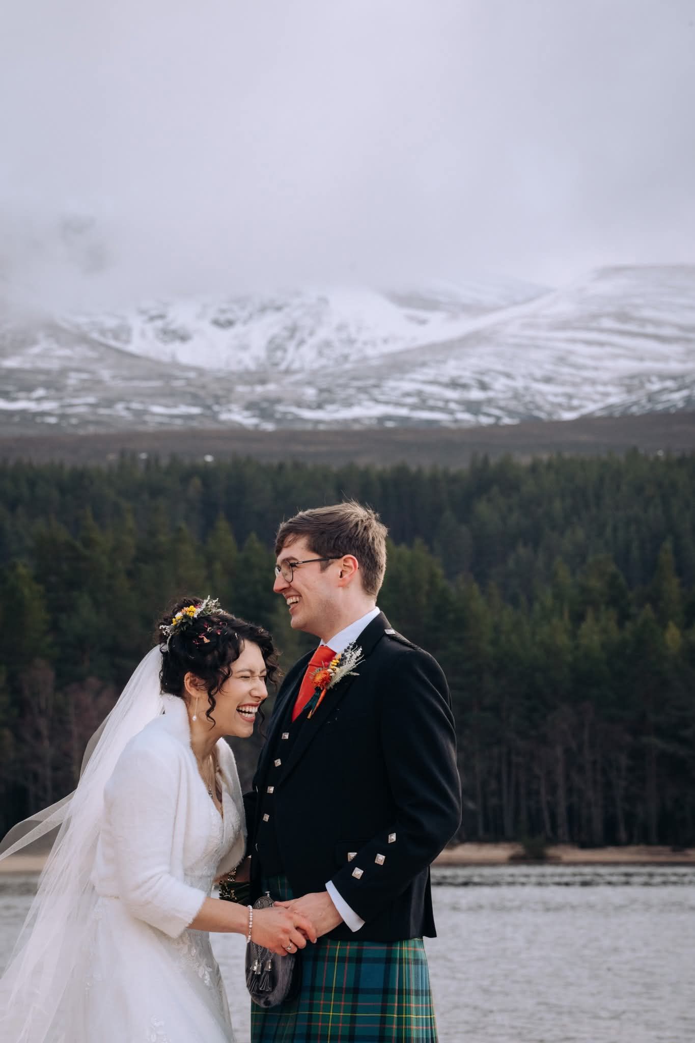 A couple shares a romantic moment at their Scottish Highland wedding with snow-capped mountains and a lake in the background.