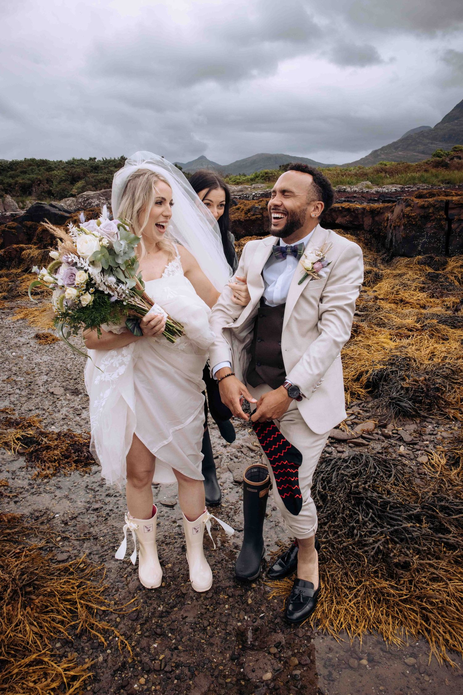 A couple shares playful moments during their coastal wedding photoshoot on rugged rocky terrain under moody skies.