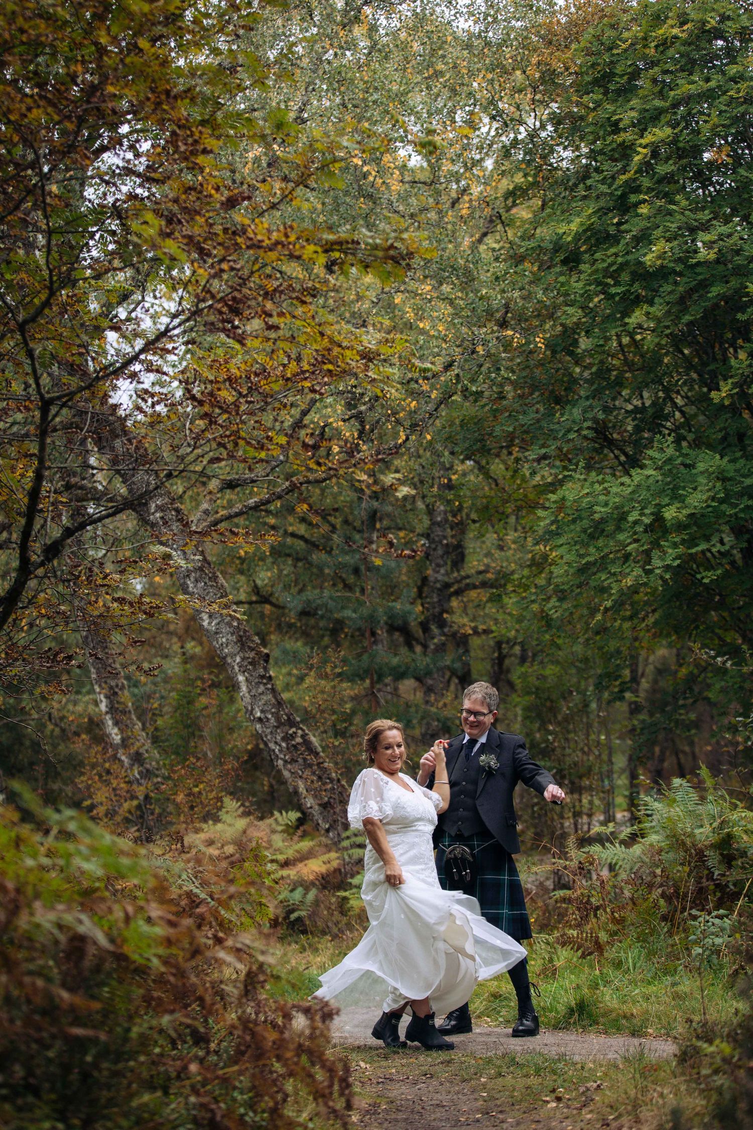 A couple in wedding attire dances playfully along a wooded autumn path surrounded by fall foliage.