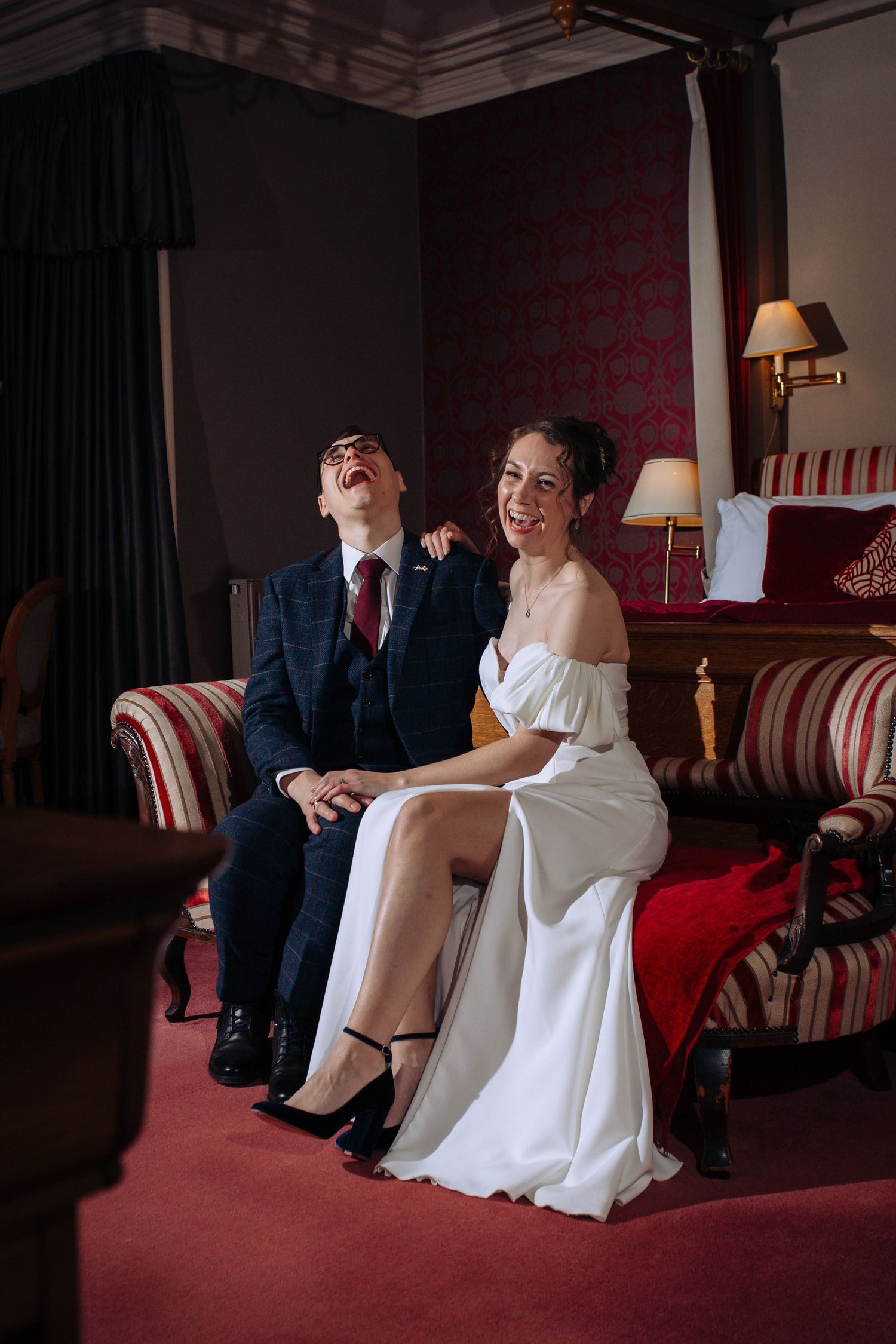 A couple shares a joyful moment while sitting on an elegant red couch in a dimly lit room with vintage decor.