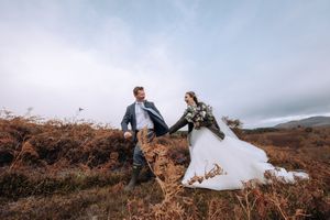 A couple in wedding attire stands on a rugged mountainside during a cloudy day with dramatic scenery.