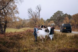 A group of people gather in a rural field during autumn with vehicles parked nearby and bare trees in the background.