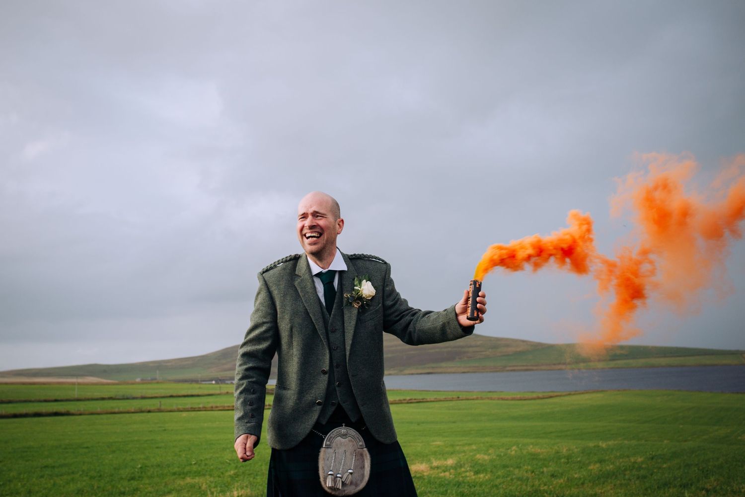 Person in suit walks across green field with orange smoke flare creating dramatic effect in cloudy weather.