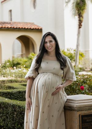 A person wearing a cream-colored dress with floral embellishments stands in front of a Mediterranean-style building.