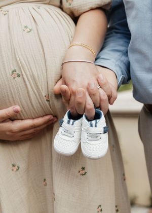 Small white baby shoes held against a pregnant belly wearing a cream colored dress.