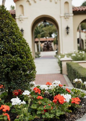 Red and white flowers bloom in a garden bed against a beige Spanish colonial building with arched windows.