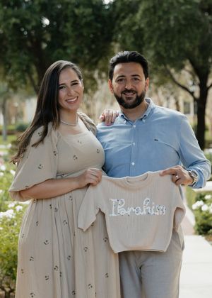 A couple poses outdoors holding a small baby outfit during a pregnancy announcement photoshoot.