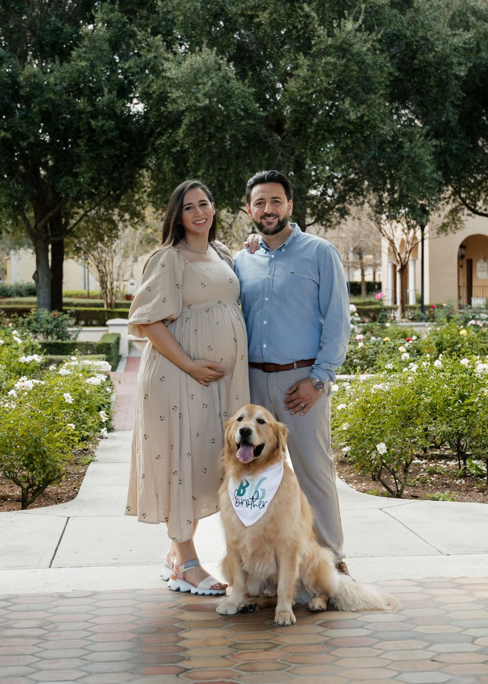 A couple stands with their Golden Retriever wearing a bandana on a brick pathway surrounded by trees and flowers.