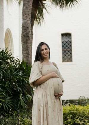 A pregnant woman in a beige floral dress poses in front of white Spanish colonial architecture and palm trees.