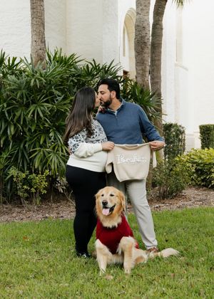 A couple shares a tender moment outdoors with their happy Golden Retriever wearing a red sweater.
