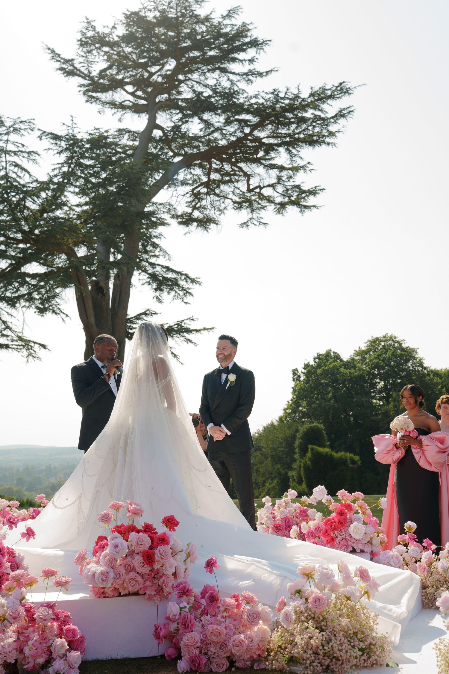 An outdoor wedding ceremony under a large tree with pink floral decorations and a long white wedding dress train.