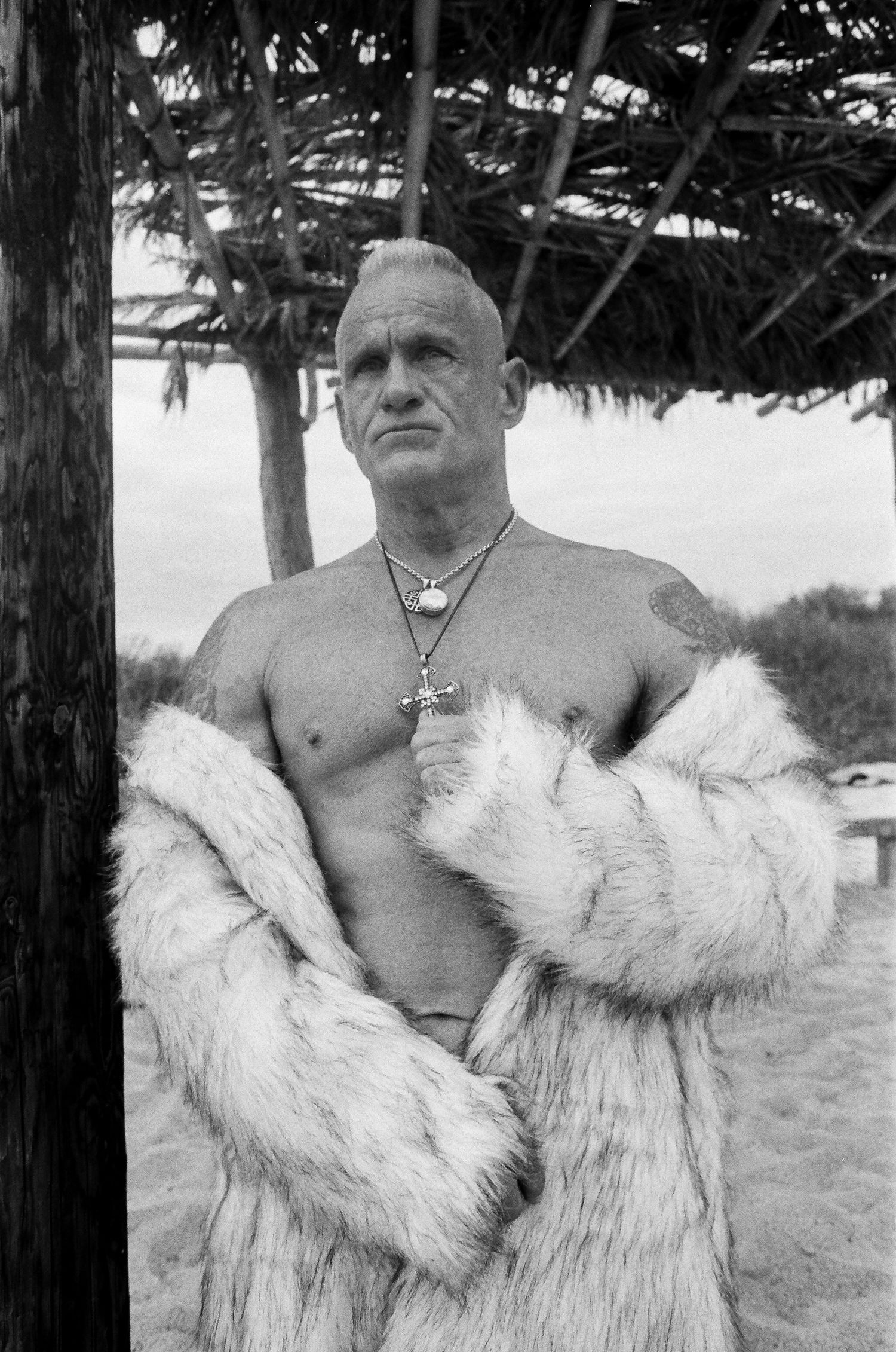 A muscular shirtless person wearing a fur coat and necklaces stands outdoors beneath tree branches.