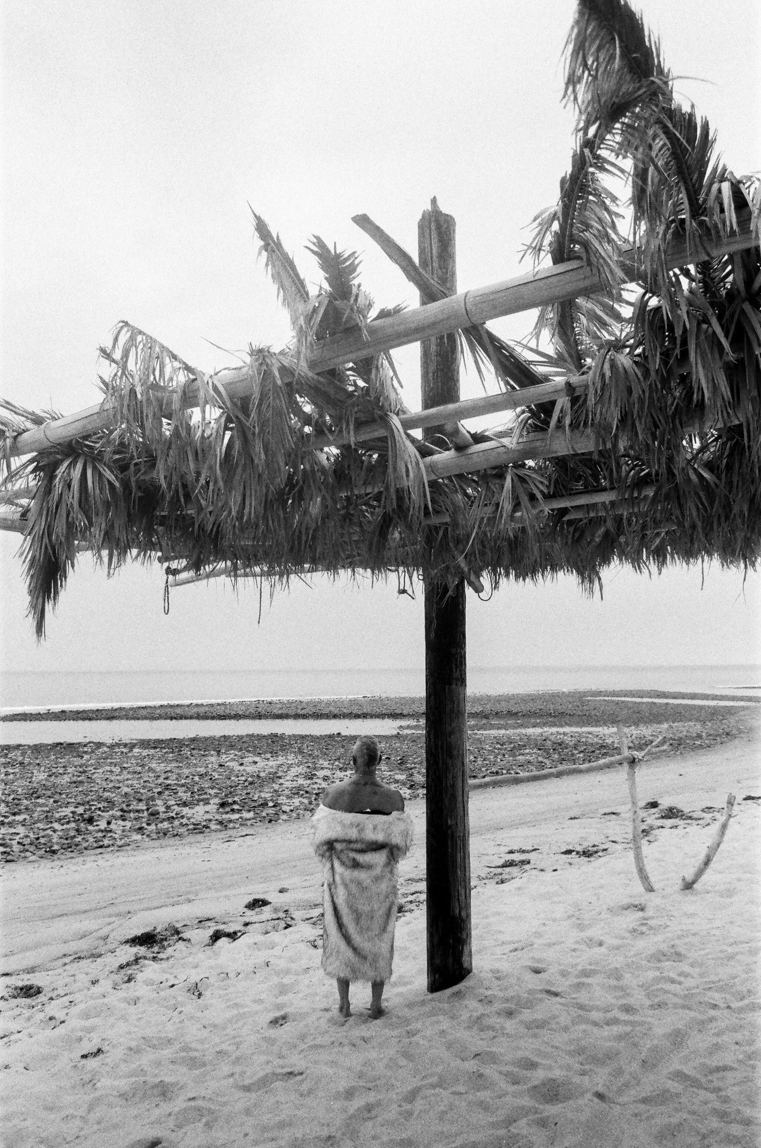Palm fronds hang from a wooden structure on a beach with figures standing beneath in black and white.