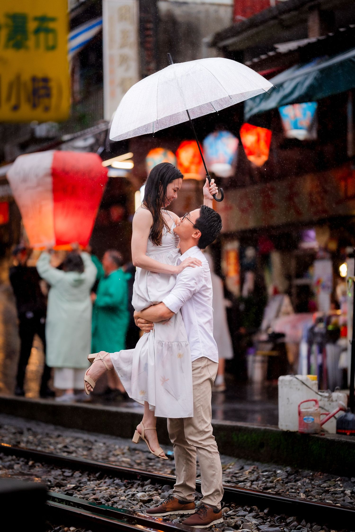 A couple in white outfits shares an intimate moment under umbrellas on a rainy street with red lanterns in the background.