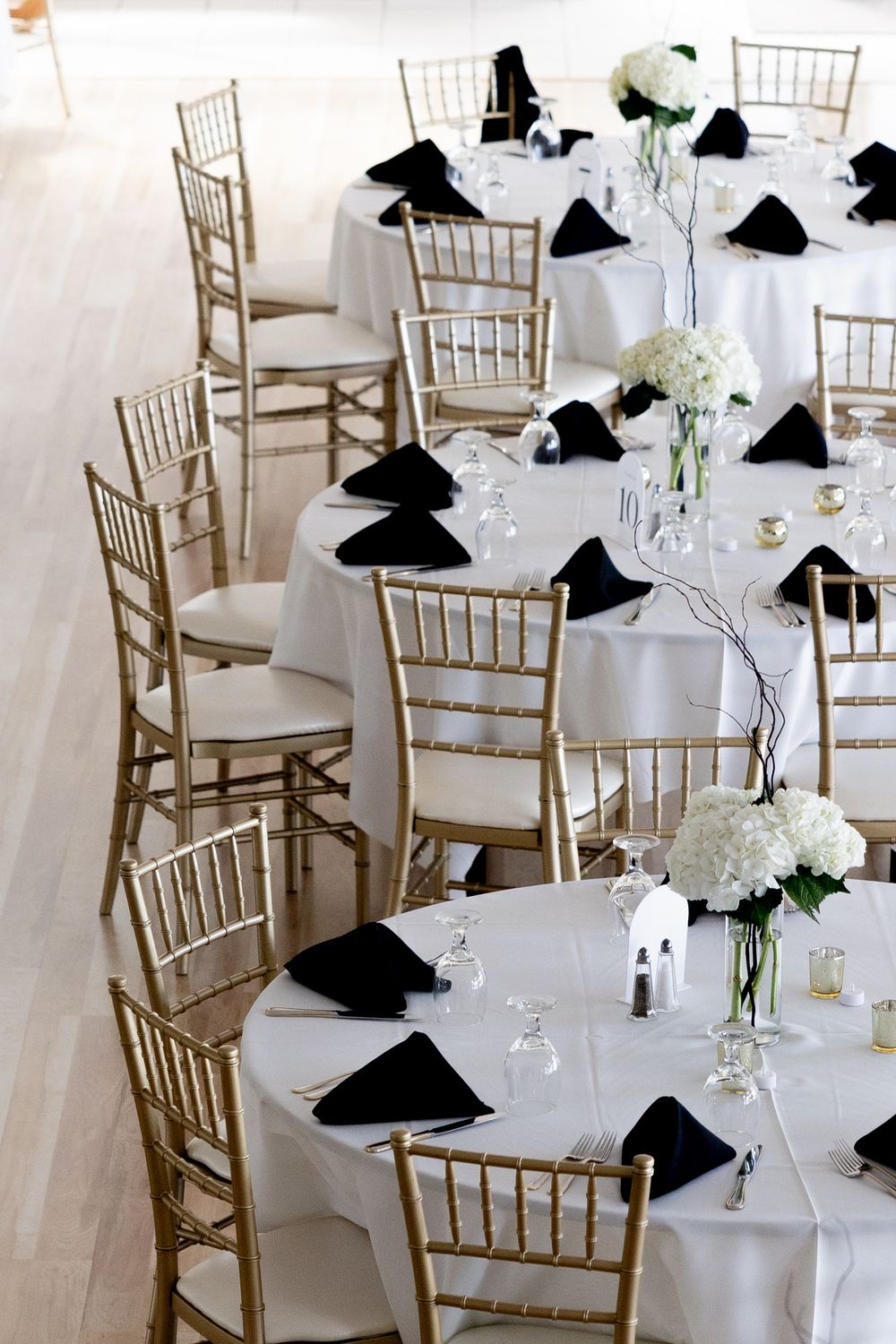 Elegant wedding reception table settings with white linens, gold chiavari chairs, and black napkins arranged in a banquet hall.