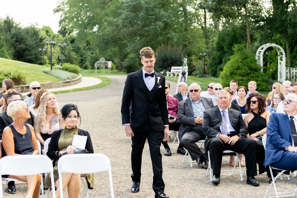 Guests seated in white chairs at an outdoor garden wedding ceremony with a scenic path and greenery in the background.