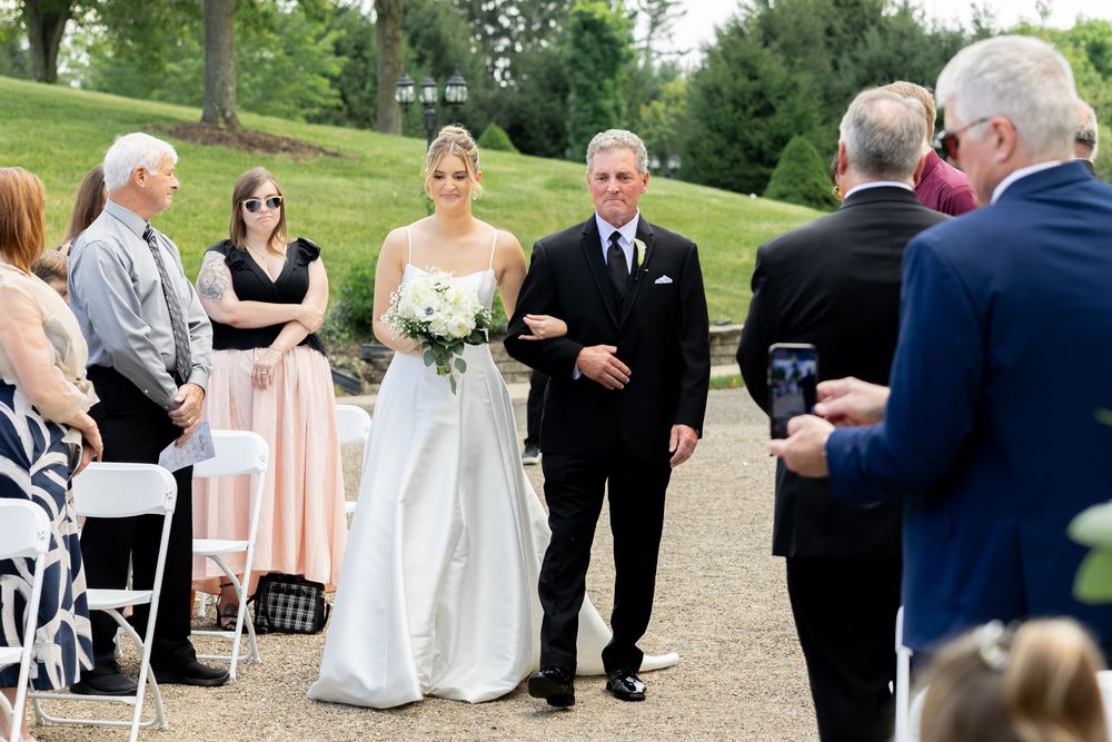 A wedding ceremony processional outdoors with guests watching the bridal party walk down a grassy path.