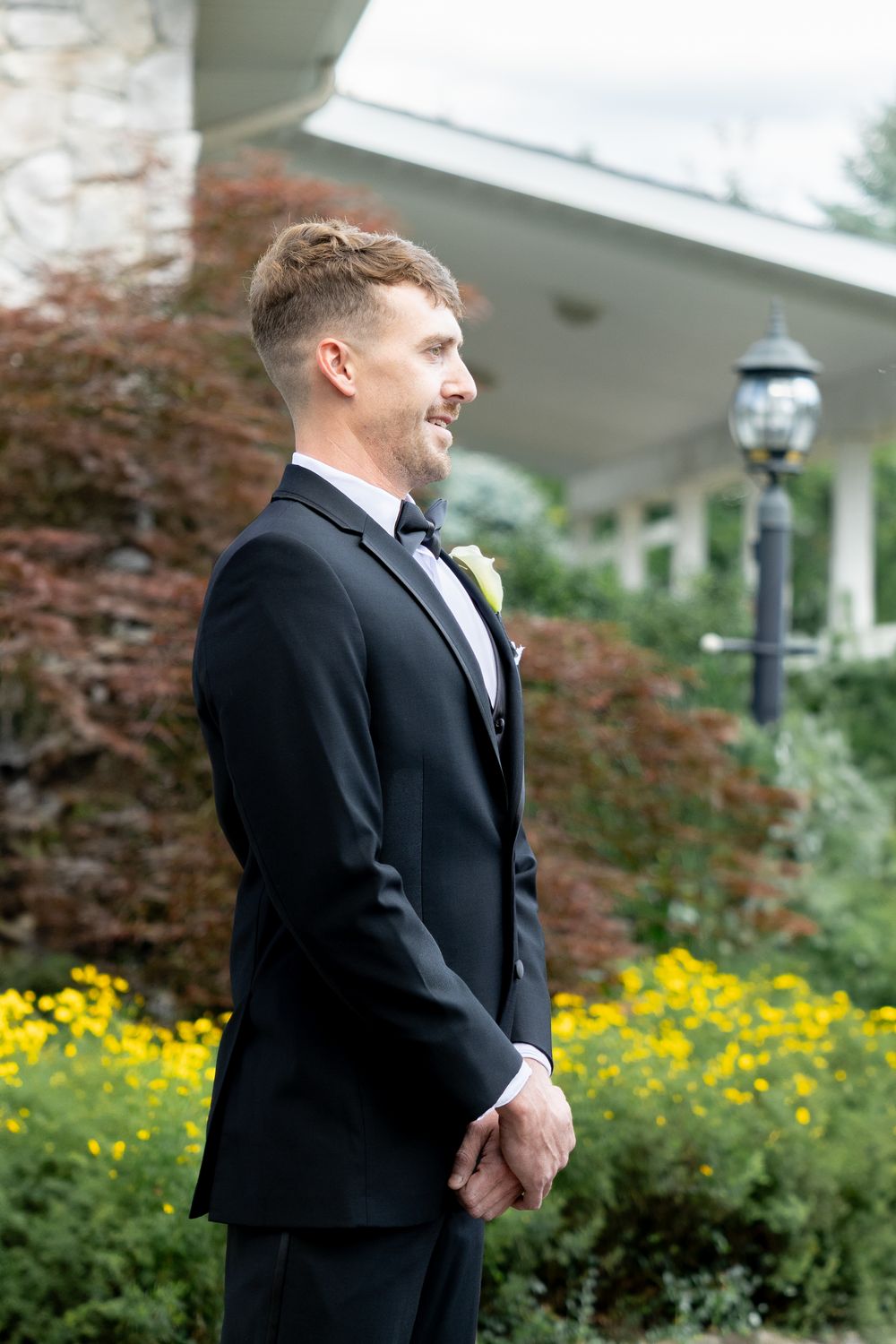 A groom in a black tuxedo stands outdoors next to yellow flowers and landscaping during a wedding photoshoot.