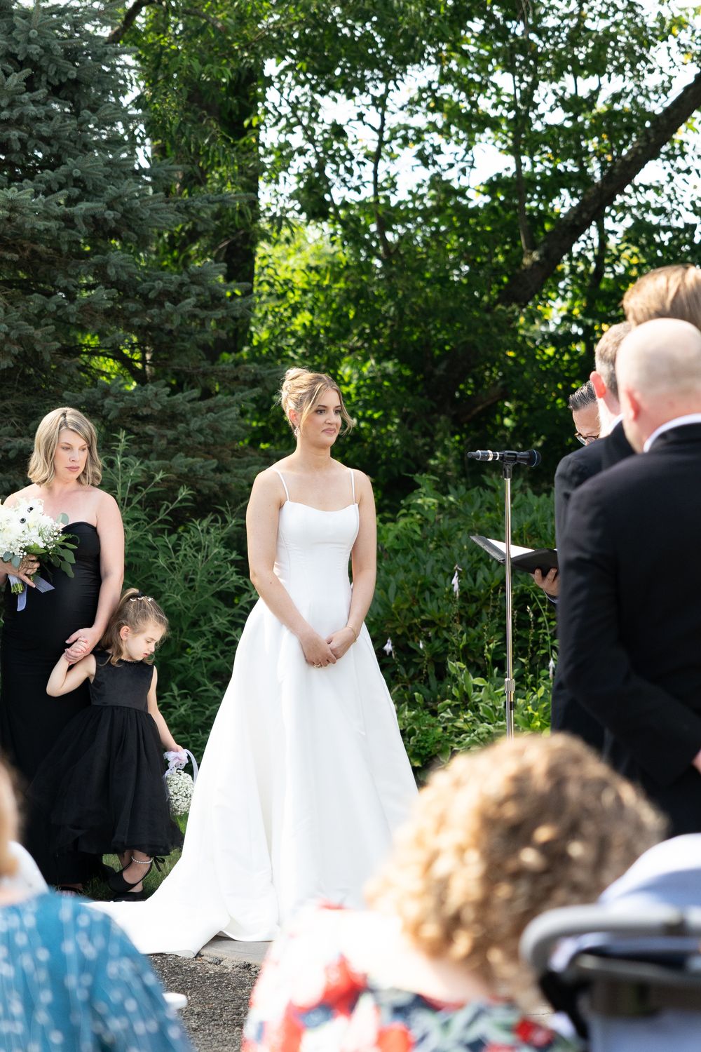 Wedding ceremony takes place outdoors with guests seated in a garden setting surrounded by lush greenery.