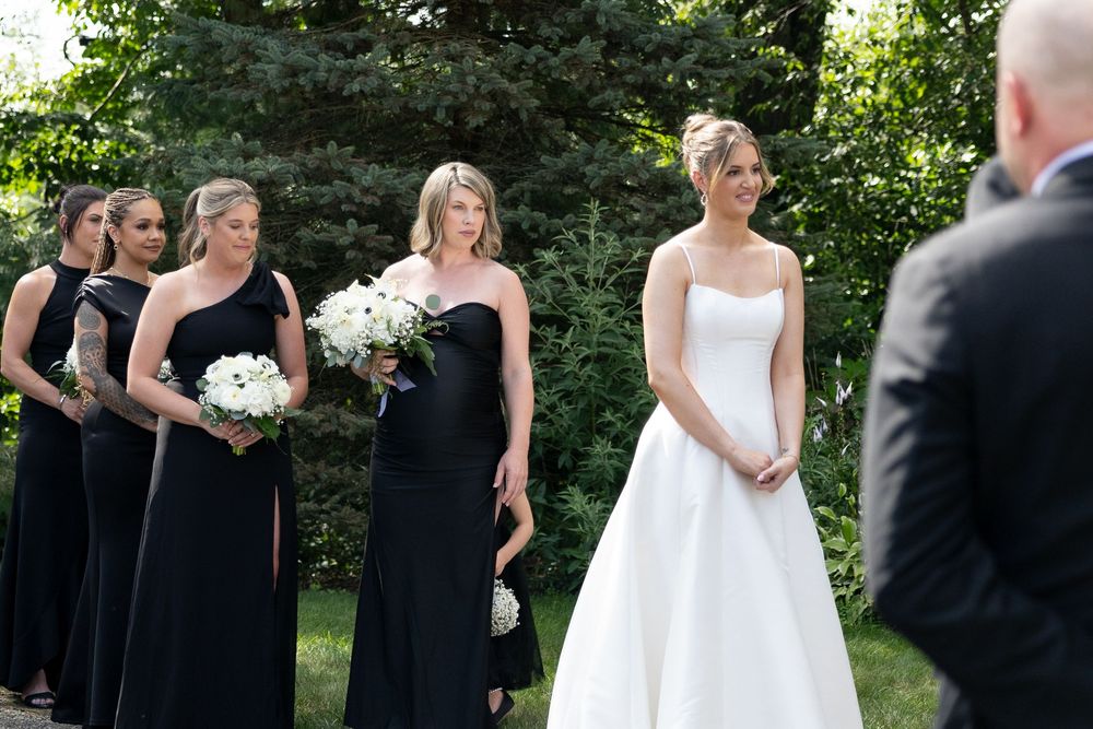 Wedding party gathers outdoors in formal attire with bridesmaids in black dresses and a bride in white.