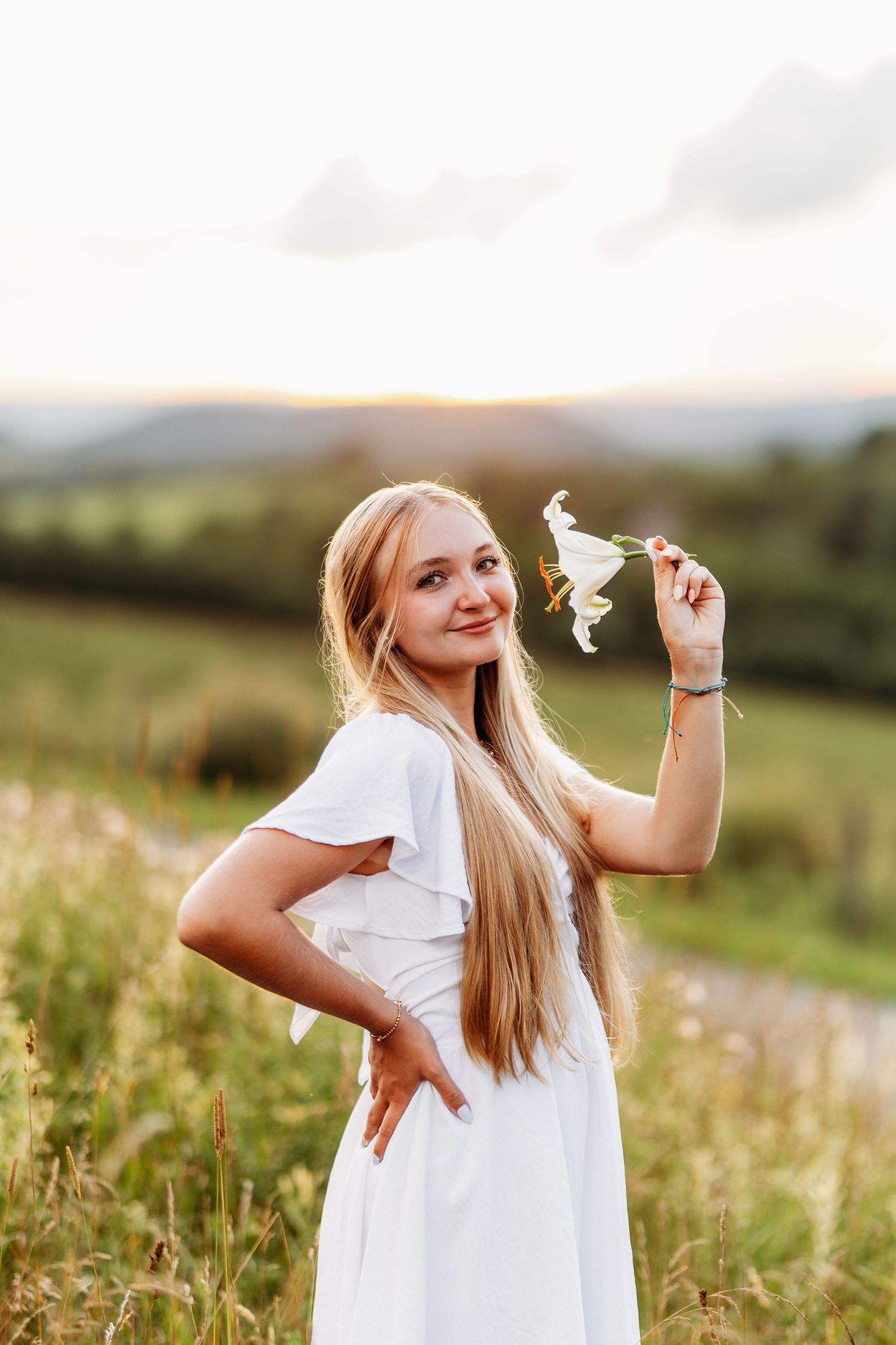 Blonde girl in white dress holding wildflower while standing in green field at golden sunset.
