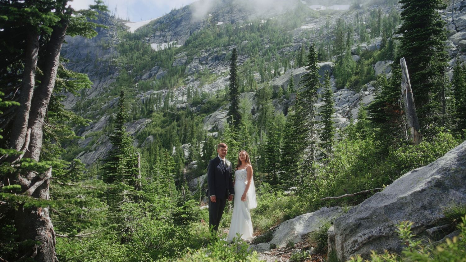 A romantic mountain wedding photoshoot with a couple embracing against a backdrop of evergreen forests and cloudy skies.