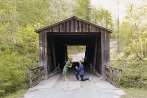 A couple shares a marriage proposal moment at the entrance of a rustic wooden covered bridge surrounded by lush green trees.