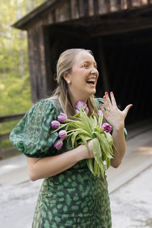 Happy woman in green floral dress laughing while holding purple tulips near a rustic wooden barn.