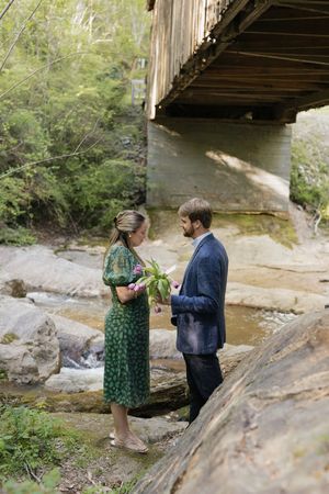 Couple stands on rocky creek bed beneath wooden covered bridge surrounded by lush green forest.