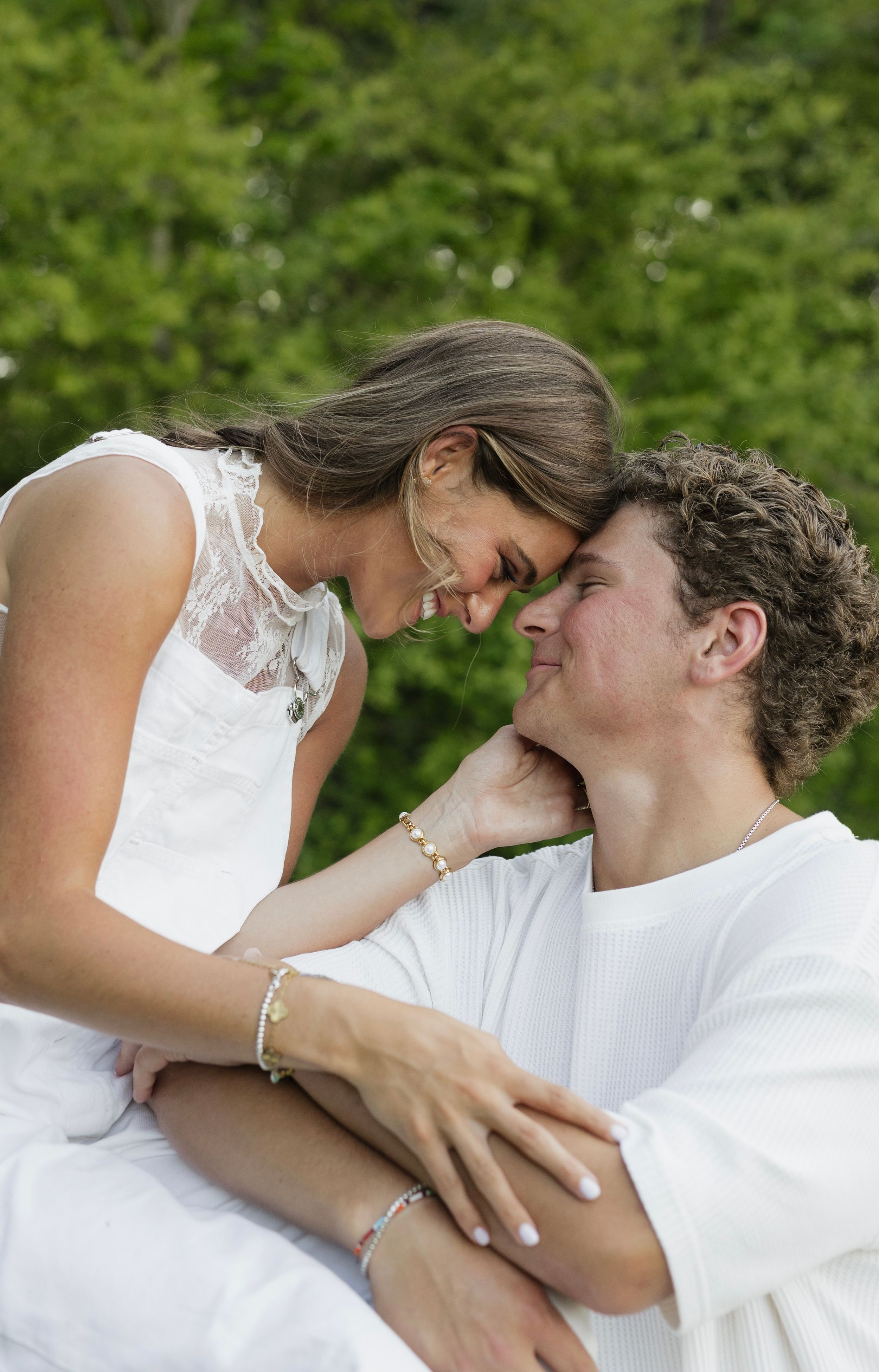 Romantic couple in white outfits sharing an intimate moment outdoors with lush green trees in the background.