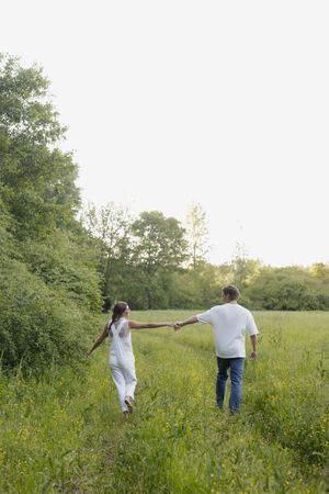 Couple holding hands walking through a lush green meadow path surrounded by trees in warm sunset light.