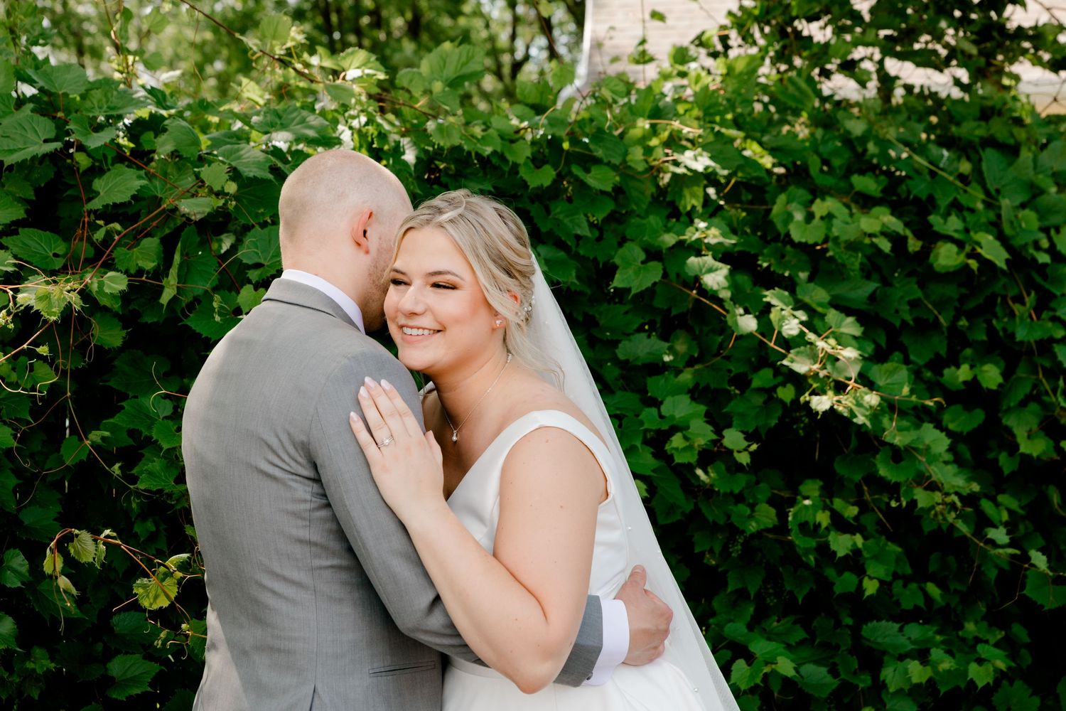 Bride in white gown and veil embraces groom in gray suit outdoors with lush green foliage backdrop.