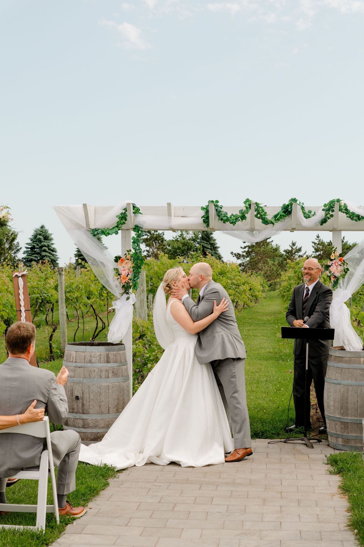 Bride and groom share first kiss at outdoor vineyard wedding ceremony under floral arch with officiant and guests.