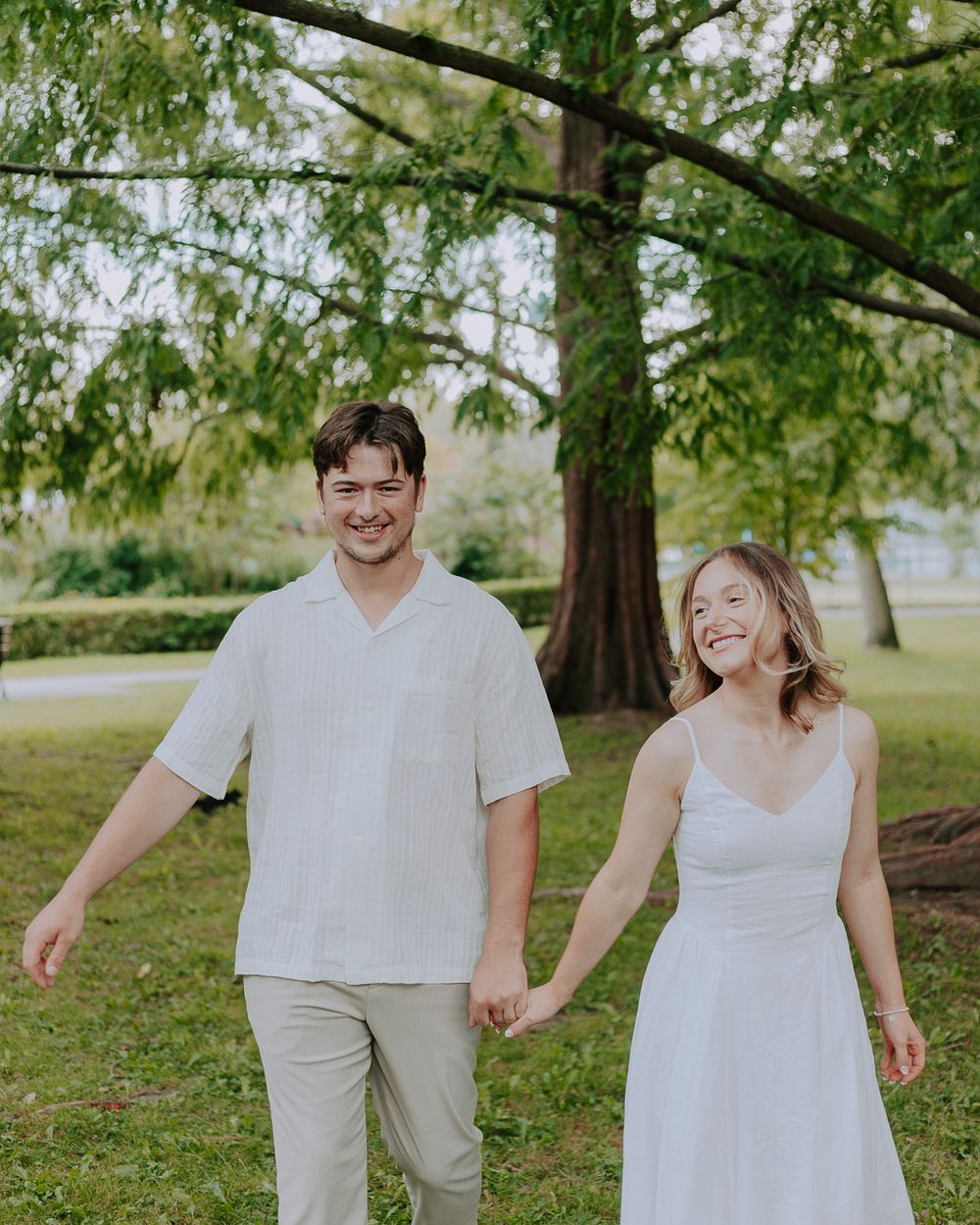 A couple in white outfits holding hands and laughing together in a park with lush green trees.