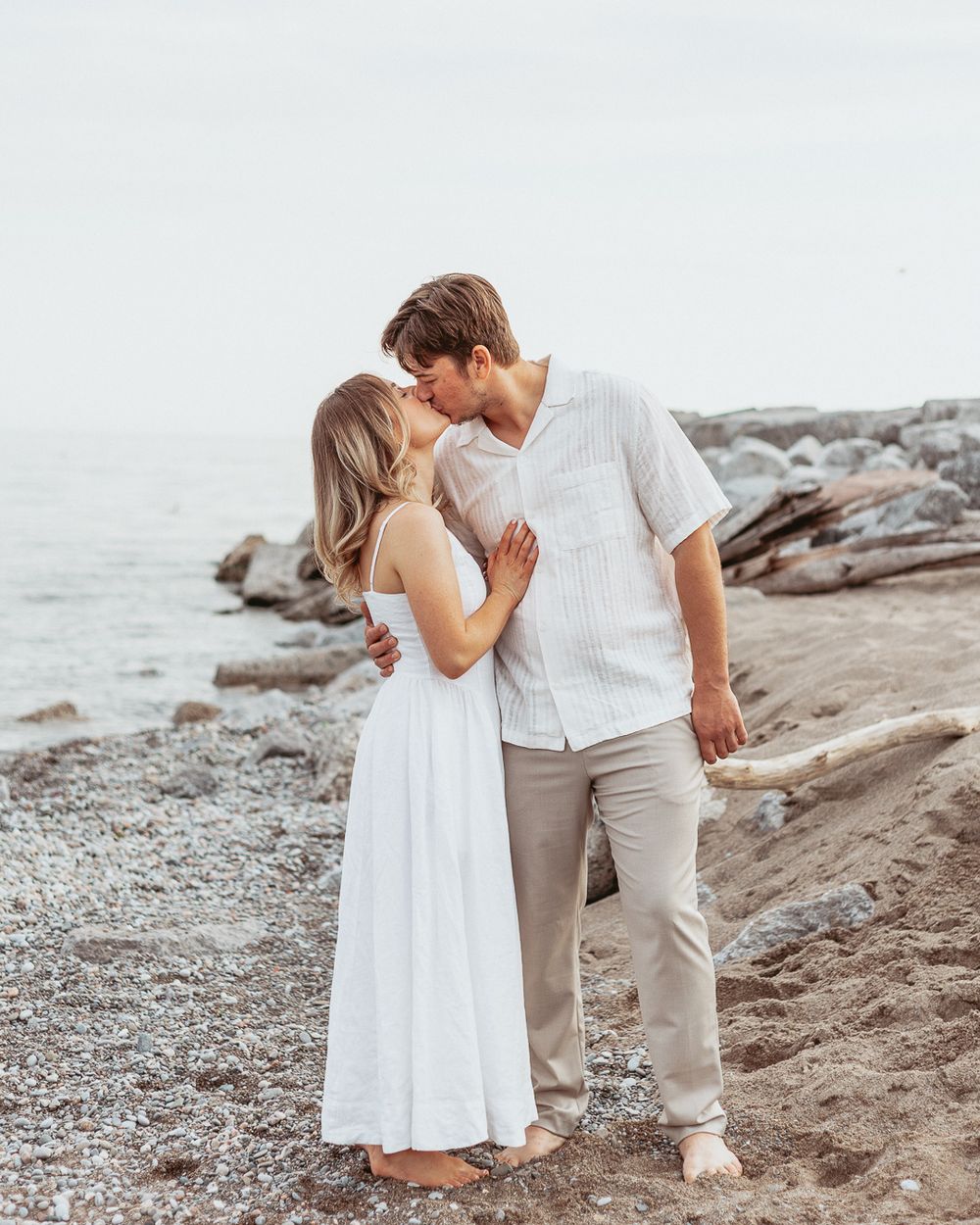 Romantic couple in white outfits sharing an intimate moment on a rocky beach.