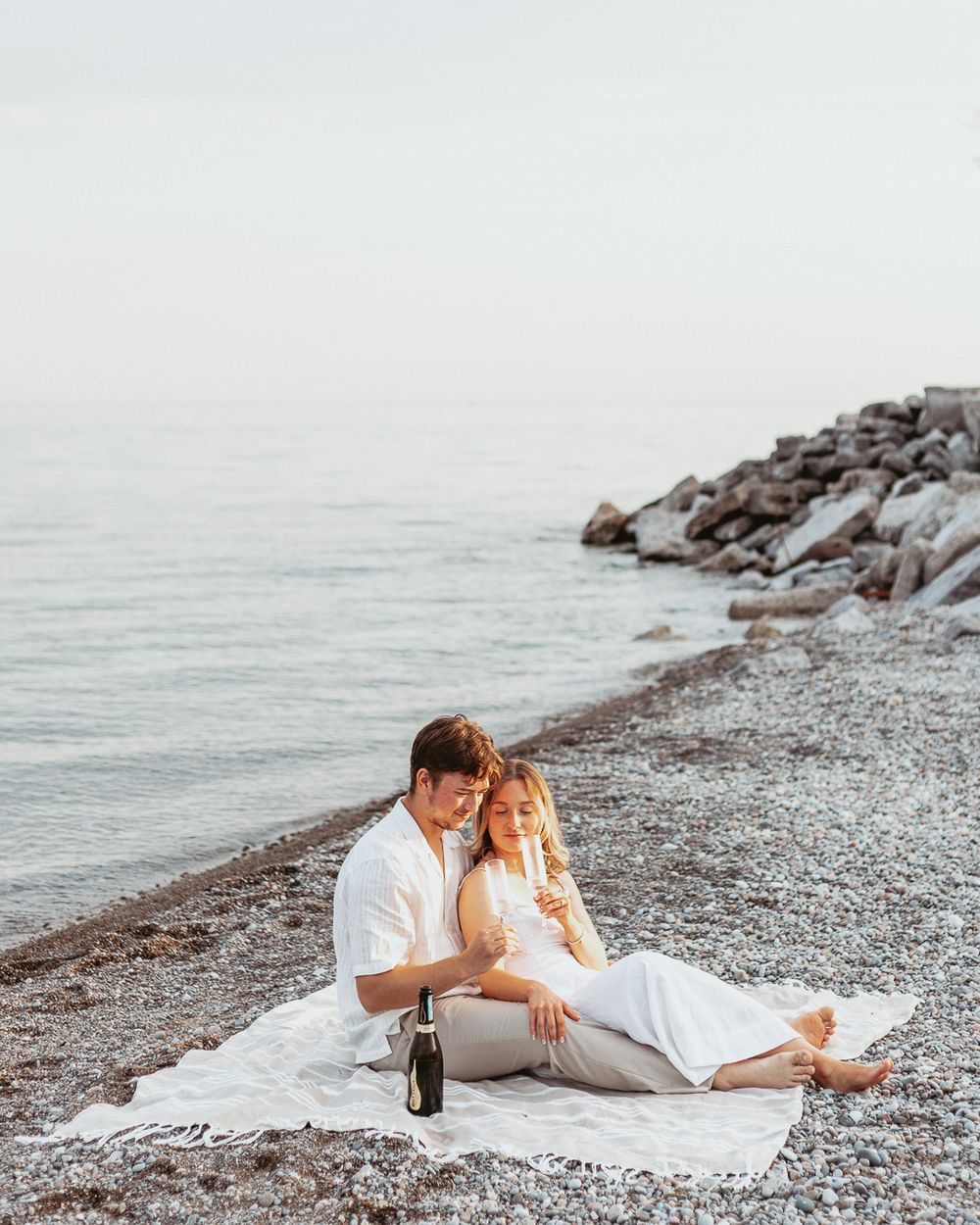Romantic couple enjoying a picnic on a rocky beach with a bottle of wine at sunset.