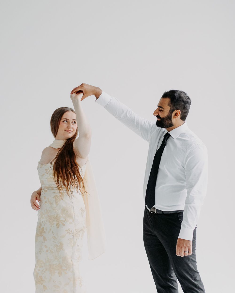 A groom in a white shirt and black tie twirls his bride in a floral wedding dress against a white background.