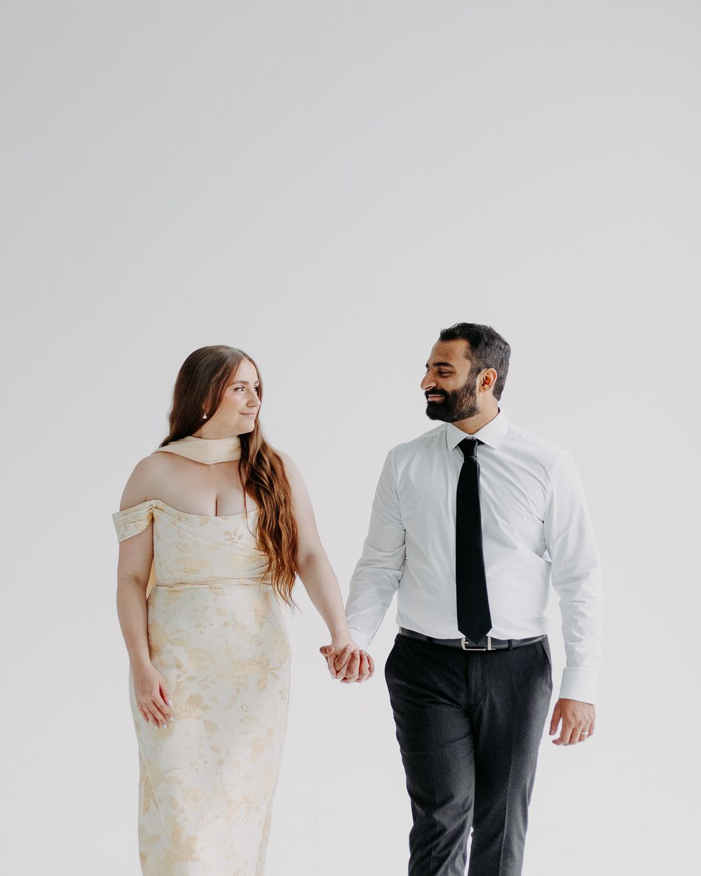 A couple holds hands and gazes at each other, woman in cream gown, man in white shirt and black tie.