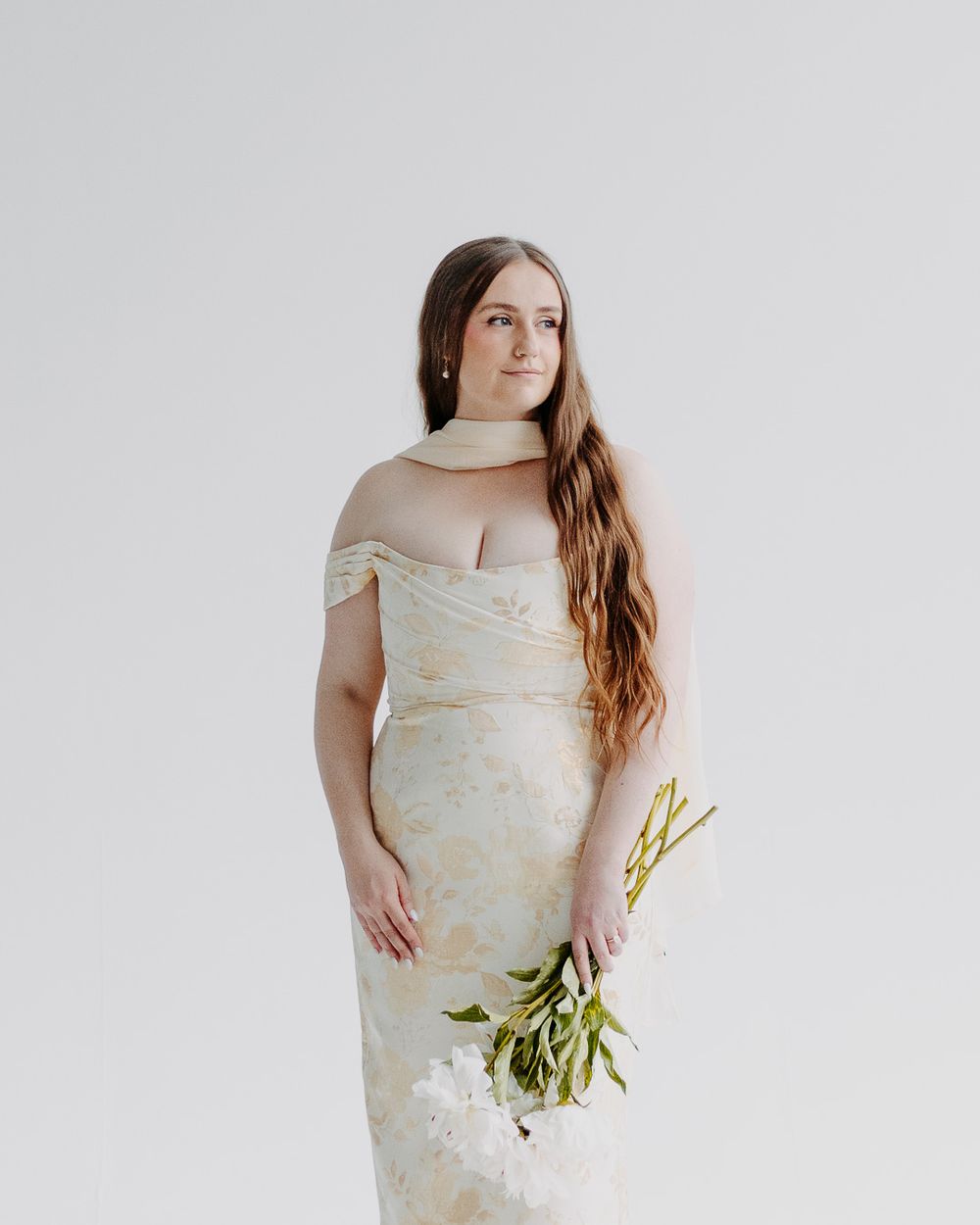 A bride in an off-shoulder floral gown holds white flowers, posing gracefully against a soft white background.